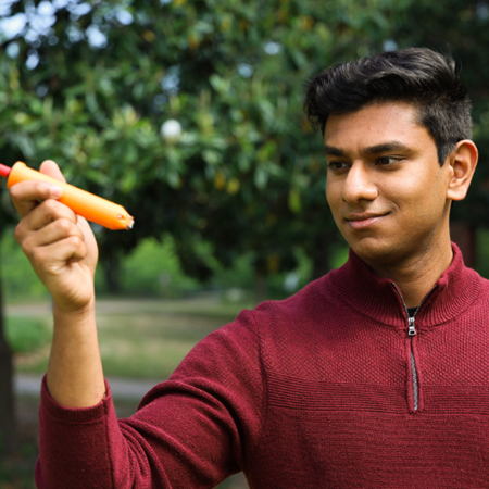 A man stands outside among trees, holding up and examining an orange pen-like device in his hand