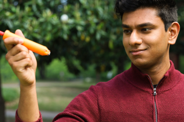 A man stands outside among trees, holding up and examining an orange pen-like device in his hand