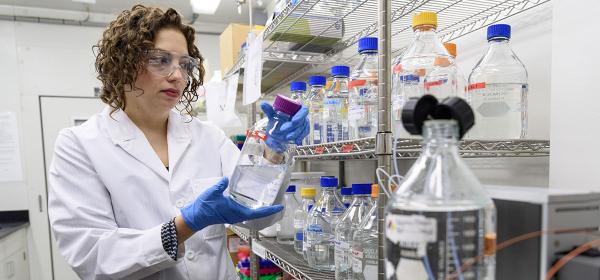 Dr. Raquel Lieberman in the lab working with glass labware and tubing, next to bottle-laden metal shelving. 